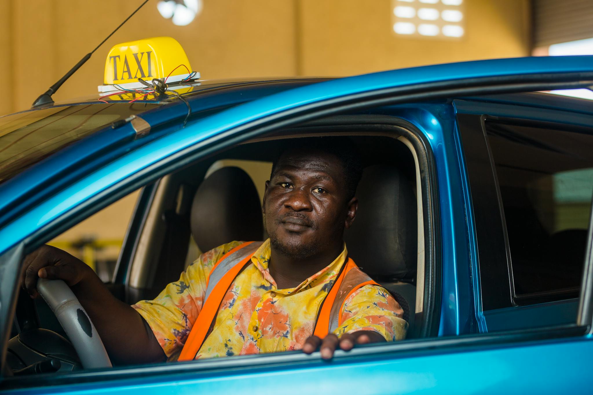 A Ghanaian taxi driver in a colorful shirt sits inside his blue vehicle, ready for passengers.